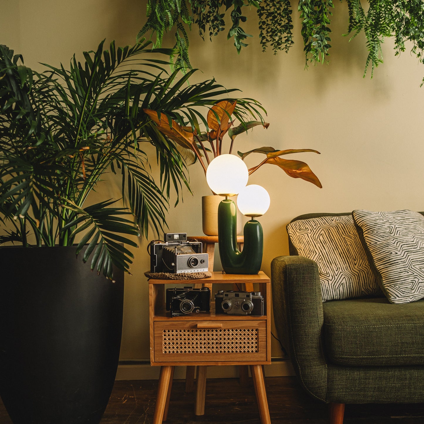 Living room with a green table lamp, sofa, wicker side table, and decorative plants.